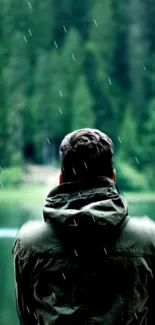 Man in jacket gazes at serene forest lake with raindrops falling.