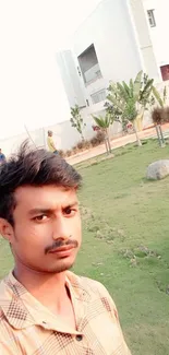 Young man takes selfie in green garden with modern building backdrop.