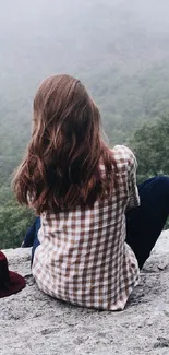 Woman in plaid shirt sitting on mountain overlooking a misty forest.