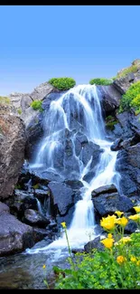 Serene mobile wallpaper of a mountain waterfall with blue skies and vibrant flowers.