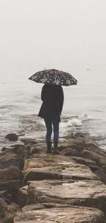 Person with umbrella standing on rocky shore by serene ocean.