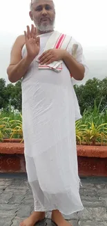 Monk standing outdoors in white attire surrounded by greenery.