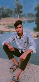 Young man sitting by a serene riverside backdrop.