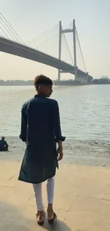 Man walking by a riverside with a large bridge in the background.