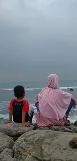 Mother and child sitting by the ocean with gray sky.