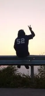 Silhouette of person at sunset holding peace sign, sitting on guard rail.