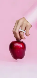 Hand with ring holding red apple on pink background.