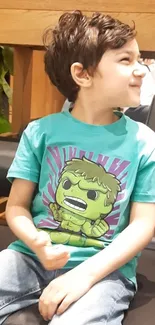 Child in turquoise T-shirt with a cartoon hero design, sitting indoors.