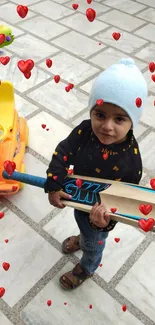 Child holding a cricket bat with hearts and a toy car on a tiled floor.