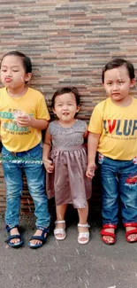 Three children standing against a brick wall in colorful attire, smiling brightly.