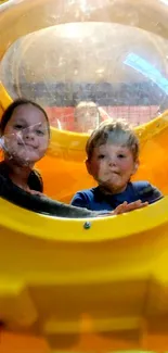 Two children smiling inside a yellow play capsule.