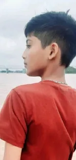 Young boy gazes over a calm river under a cloudy sky.