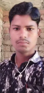 Man in a patterned shirt standing against a rustic brick wall.