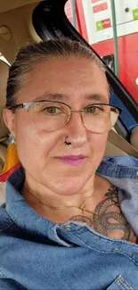 Woman sitting in car with denim shirt and glasses, showing tattoos and a calm expression.