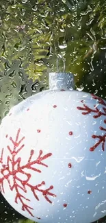 White Christmas ornament with red snowflakes behind raindrop-covered glass.