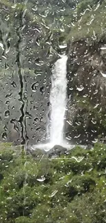 Rainy window view of a waterfall surrounded by lush green scenery.