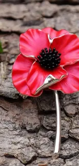 Red poppy brooch on rough bark background with green leaves.