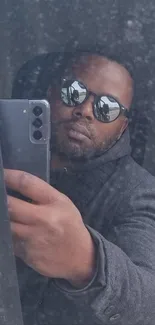 Man taking a selfie in a window reflection with sunglasses and smartphone.