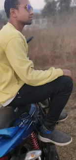 Young man in yellow shirt sitting on a blue motorcycle in nature.