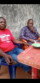 Two men relaxing outdoors with food and drinks by a wall.