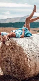 Woman in blue dress relaxing on hay bale in open field.