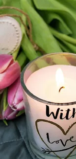 Candle with 'With Love' text surrounded by pink tulips on a quilted surface.