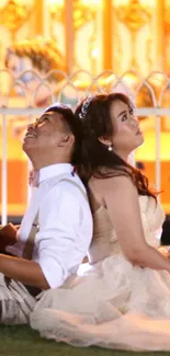 A couple in wedding attire sits back-to-back under golden evening lights.