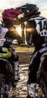 Two dirt bike riders share a moment under a vibrant sunset on a muddy track.