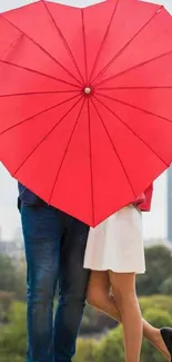 Couple under heart-shaped red umbrella with city landscape.