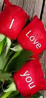 Three red roses with 'I love you' text on wooden background.