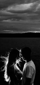 Silhouette of a couple by a lake under dramatic clouds in black and white.