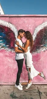 Couple embraces in front of colorful street art with pink wings.