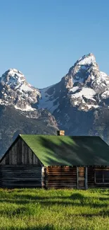 Rustic cabin in front of snow-capped mountains and green grass.