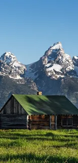 Rustic cabin with green roof set against snowy mountains.