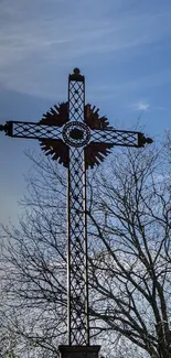 Silhouette of a cross against a blue sky with bare tree branches.