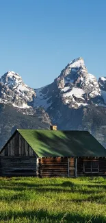 Rustic cabin in green field with snowy mountains in the background.