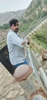 Man stands by a railing, overlooking a scenic hilltop view with greenery.