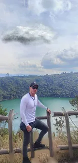 A person stands by a wooden fence overlooking a serene lake with hills and a cloudy sky.