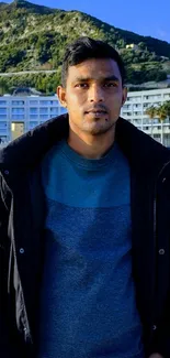 Man standing by a scenic ocean view with mountains and modern buildings.