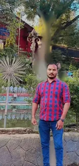 A man stands in a scenic park with a red building and lush greenery in the background.