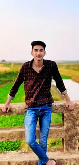 Young man on bridge with green fields and river backdrop.