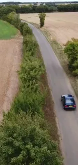 Aerial view of a car driving along a scenic countryside road with fields and trees.