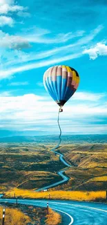 Hot air balloon over scenic winding road in golden fields and blue sky.