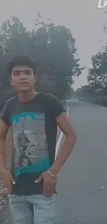 Young man standing on a scenic roadside with lush trees and a peaceful road.