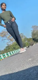 Woman walking along a sunny road with blue sky and green landscape.