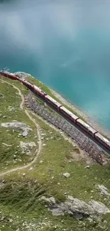 Red train passing by a turquoise lake with green hills.