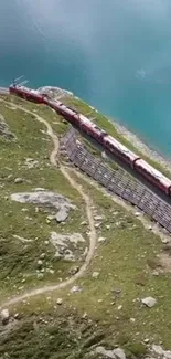 A red train passes by a blue lake on rocky green terrain.