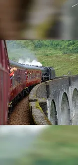 Steam train crossing a stone bridge with lush greenery in the background.