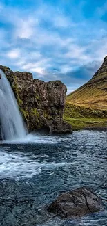 Waterfall flowing into a river beside a towering mountain under blue skies.