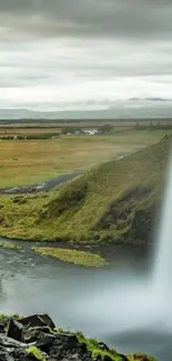 Waterfall cascading beside a lush green field under a cloudy sky.